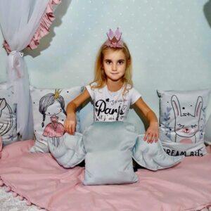 Cute girl with a pink crown sitting on decorative cushions in a cozy room.