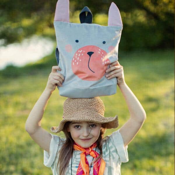 Child holding a bunny-themed kids backpack outdoors in a park setting.