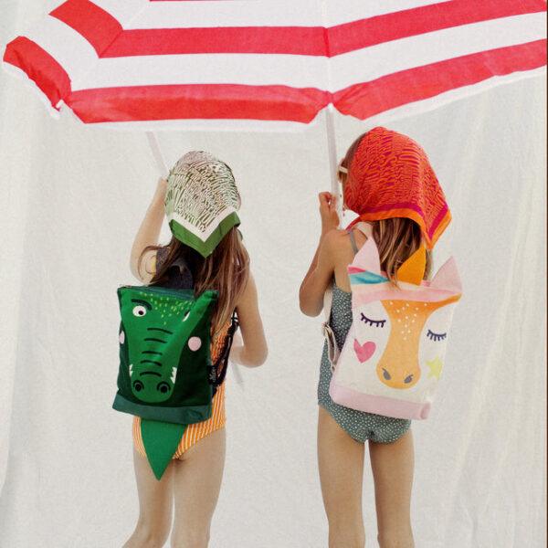 Kids backpacks with crocodile and unicorn designs under a beach umbrella.