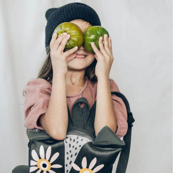 Child with a backpack holding two green apples over her eyes.