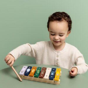 Young child playing a wooden xylophone with a mallet on a green background.